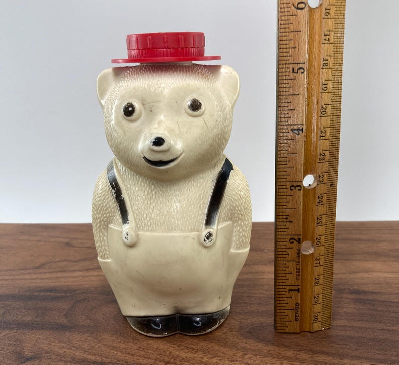 May include: A vintage bear-shaped honey bottle with a red cap. The bear is off-white with black accents on the eyes, nose, and suspenders. The bottle is approximately 5 inches tall, as measured by the wooden ruler.