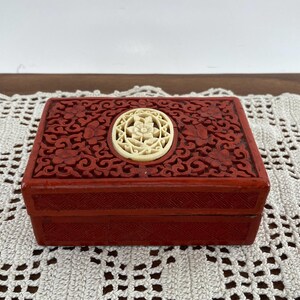 May include: A rectangular, red lacquered box with intricate floral carvings and a central, circular ivory-colored medallion. The box features a detailed, carved design on the lid and sides, showcasing a traditional aesthetic. The box is displayed on a white lace tablecloth.