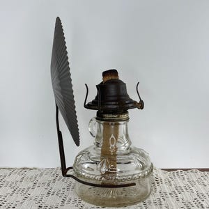 May include: Antique clear glass oil lamp with a decorative metal shade. The lamp has a round base with embossed floral patterns and a metal top. The shade is a dark gray, fan-shaped piece attached to the lamp.