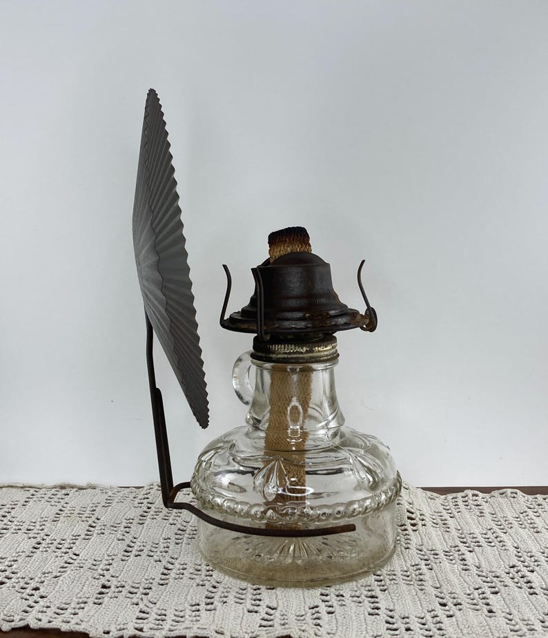 May include: Antique clear glass oil lamp with a decorative metal shade. The lamp has a round base with embossed floral patterns and a metal top. The shade is a dark gray, fan-shaped piece attached to the lamp.