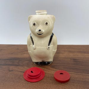 May include: A vintage cream-colored bear-shaped syrup dispenser with black accents and a red lid. The bear has black suspenders and feet. Two red lids are next to the dispenser. This collectible item is a charming addition to any kitchen.