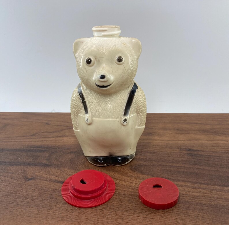 May include: A vintage cream-colored bear-shaped syrup dispenser with black accents and a red lid. The bear has black suspenders and feet. Two red lids are next to the dispenser. This collectible item is a charming addition to any kitchen.