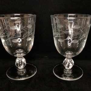 May include: Two clear, stemmed wine glasses with etched floral designs. The glasses have a rounded bowl, a decorative stem, and a circular base. The etched design features leaf-like shapes and thin lines. The glasses are displayed against a dark background.