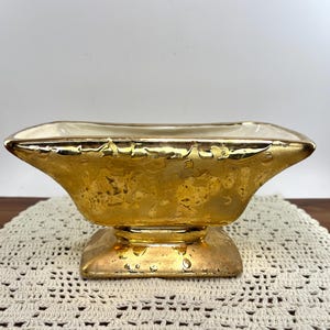 May include: A rectangular ceramic planter featuring a metallic gold exterior finish with a textured, mottled appearance. The vessel has a wide, flared rim and sits on a square pedestal base, with a smooth, off-white interior surface visible.