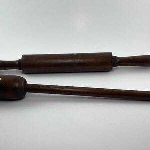 May include: Two dark brown wooden kitchen tools: a rolling pin and a muddler. The rolling pin is cylindrical with rounded handles. The muddler has a cylindrical head and a long, thin handle. Both tools are on a white background.