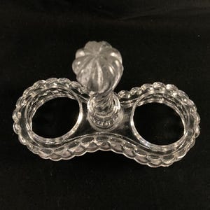 May include: A clear glass salt and pepper shaker holder with a decorative knob on top. The holder has two circular openings for the shakers.