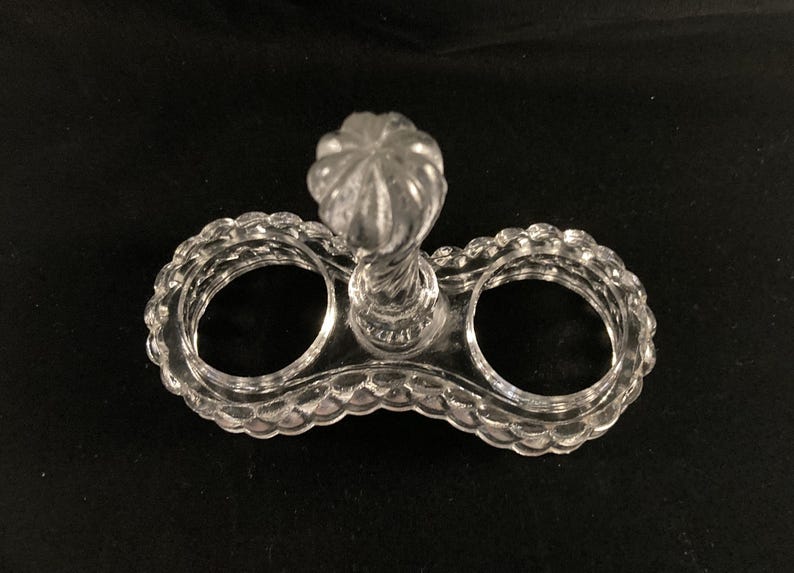 May include: A clear glass salt and pepper shaker holder with a decorative knob on top. The holder has two circular openings for the shakers.