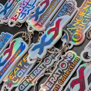 Puede incluir: Una colección de llaveros iridiscentes con varios diseños y texto. Los llaveros presentan una variedad de colores, incluyendo azul, rojo y plateado. El texto incluye frases como "Phantom Force" y "Ancient Origins". Cada llavero tiene un anillo plateado.