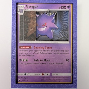 Op de afbeelding: Een ingelijste Pokémon-kaart met Gengar, een paars spookachtig personage. De kaart zit in een paarse lijst. De tekst op de kaart bevat "Gnawing Curse" en "Fade to Black". De kaart is 149.86 cm hoog en weegt 40.5 kg.