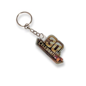Op de afbeelding: Een metalen sleutelhanger met een platte, hoekige hanger met een goud- en zwartverloop. De hanger toont de tekst 30th CELEBRATION met een klein rood en wit cirkelvormig icoon dat de letter O vervangt. Inclusief een standaard sleutelring.