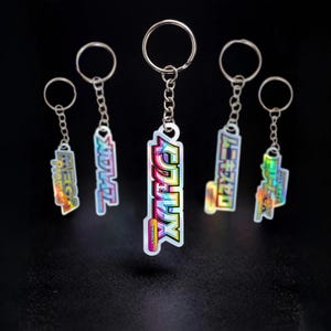 May include: Five keyrings with silver chains and rings. Each keyring features a rectangular, holographic design with Japanese text in a colourful, gradient font. The keyrings are displayed against a dark background.