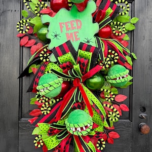 May include: A Halloween-themed door wreath with a green, red, and black color scheme. The wreath features a green wooden sign that says "FEED ME" with spider accents. It includes green, red, and black ribbons, green orbs, and decorative green and red leaves.