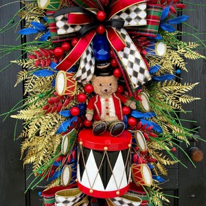 Puede incluir: Una corona navideña festiva con un oso de peluche vestido de tamborilero, sentado sobre un tambor rojo y negro. La corona incluye follaje rojo, verde y dorado, bayas rojas y tambores decorativos, acentuados con cintas a cuadros y arlequín.