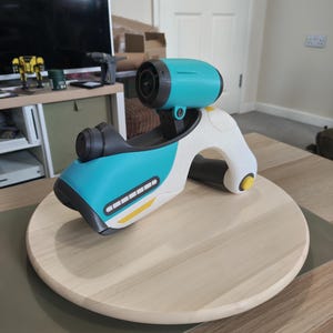 May include: A futuristic toy blaster featuring a white, teal, and dark grey colour scheme. The device has a top-mounted teal cylindrical scope, a dark grey handle, and a yellow button on the side. The body includes a series of white rectangular accents.