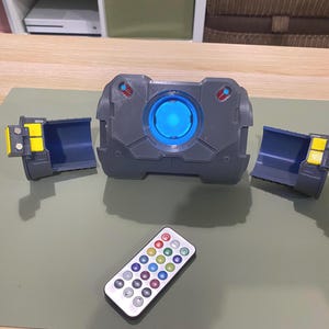 Op de afbeelding: Een grijs elektronisch apparaat met een blauw licht in het midden, twee afneembare zijstukken en een afstandsbediening met gekleurde knoppen. Het apparaat staat op een groene ondergrond.