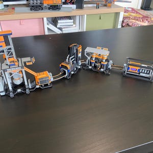Könnte beinhalten: Eine Reihe miteinander verbundener, miniaturisierter Industriemodelle in Orange, Grau und Schwarz. Die Modelle scheinen Teil eines größeren Systems zu sein, wobei verschiedene Komponenten miteinander verbunden sind. Die Strukturen stehen auf einer dunklen Oberfläche.