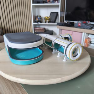 以下が含まれることがあります： 3Dプリントされたモジュール式宇宙ステーションのおもちゃ。白とターコイズブルーのベースにダークグレーの天板があり、ターコイズブルーの窓、黄色の手すり、白と黄色の円形ドッキングポートを備えた白い円筒形のトンネルセクションが接続されています。