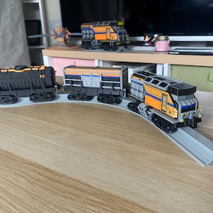 Könnte beinhalten: Ein Spielzeug-Zugset mit einer schwarzen, orangefarbenen und grauen Lokomotive und Waggons auf einer grauen Strecke. Die Züge umfassen einen Tankwagen und einen Güterwagen. Der Zug steht auf einer Holzoberfläche.