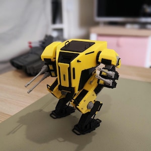 Puede incluir: Un robot de juguete amarillo y negro con dos cañones y dos armas más pequeñas. El robot tiene un diseño cuadrado con piernas y brazos articulados. Un tanque de juguete gris es visible en el fondo.