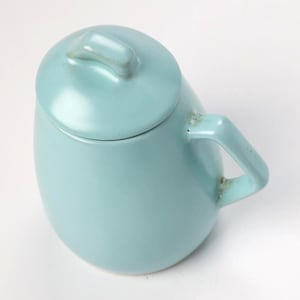 Op de afbeelding: Een lichtblauwe keramische mok met een bijpassende deksel. De mok heeft een afgeronde vorm met een handvat en een kleine knop op het deksel voor gemakkelijk verwijderen. De mok staat op een witte achtergrond.