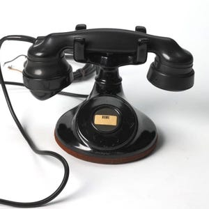 May include: A vintage black rotary telephone with a handset resting on a cradle. The base features a circular center with a small rectangular label printed with the word ROME. A long black cord extends from the handset to the base of the unit.