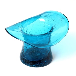May include: A turquoise glass vase with a unique, curved design. The vase has a wide, flared top and a textured, crackled surface. The base is a darker shade of turquoise. The vase is a decorative item.