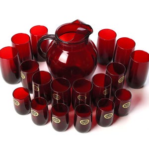 May include: A set of deep red glass drinkware consisting of one large pitcher with a handle and spout, accompanied by sixteen matching tumblers. Each tumbler features a small circular gold sticker on the exterior surface of the glass.