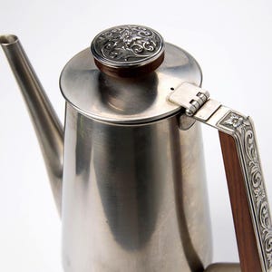 Może przedstawiać: Srebrny dzbanek do kawy z długim dziobkiem i ozdobnym uchwytem. Uchwyt wykonany jest z drewna i ma bogate srebrne detale. Pokrywa ma srebrny kwiatowy wzór. Dzbanek przeznaczony jest do parzenia kawy.