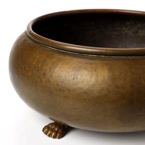 May include: A round, bronze-colored metal bowl featuring a hammered texture on its exterior surface. The bowl is supported by three small, detailed feet shaped like animal paws, and it has a smooth, rounded rim with a deep, open interior.