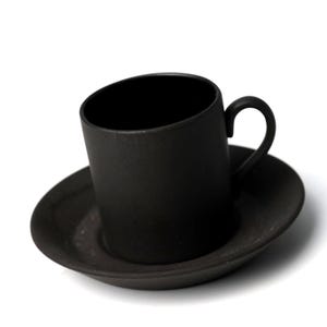 May include: A matte black ceramic teacup and saucer set. The cup has a curved handle and sits on a matching saucer. The set is a solid, dark color, and the design is minimalist and modern. The cup and saucer are made of ceramic.