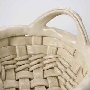 May include: Cream-colored ceramic basket with a woven design. The basket features a curved handle and a textured surface, showcasing a handcrafted aesthetic. The basket's design is reminiscent of traditional woven baskets.