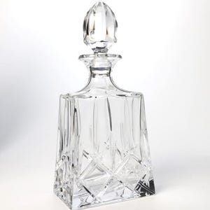 May include: A clear glass decanter with a square base and a faceted, pointed stopper. The body features a geometric diamond-cut pattern on the lower section, transitioning into vertical fluted panels that extend upward toward the neck.