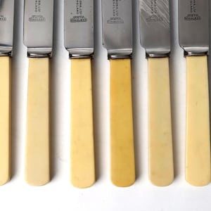 May include: Six vintage knives with stainless steel blades and cream-colored handles. The blades have a rectangular shape and are attached to the handles with a silver-colored metal. The handles are a light cream color.