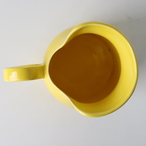 May include: Overhead view of a bright yellow ceramic pitcher with a rounded body and a curved handle. The interior of the pitcher is a slightly darker shade of yellow. The pitcher has a spout for easy pouring. The background is plain white.