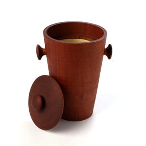 May include: A cylindrical wooden ice bucket with a rich brown grain, featuring two small side handles and a matching circular lid placed beside it. The interior of the bucket is lined with a smooth, light-colored material, contrasting with the exterior.
