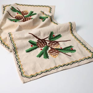 May include: A cream-colored fabric table runner featuring embroidered pinecones, green pine needles, and small red berries. The edges are finished with a decorative wavy trim in black, green, and yellow thread, creating a textured border.