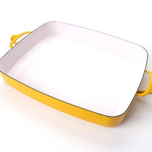 May include: A rectangular baking dish with a bright yellow exterior and a smooth white interior. The dish features two integrated yellow handles on opposite sides and a dark rim along the top edge of the vessel, providing a sturdy construction.
