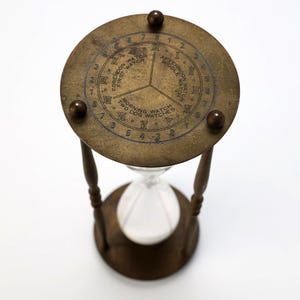May include: An antique brass hourglass with a circular top featuring Roman numerals and the text "Forenoon Watch", "Afternoon Watch", and "Morning Watch". The hourglass has a clear glass bulb and wooden supports. The design is vintage.