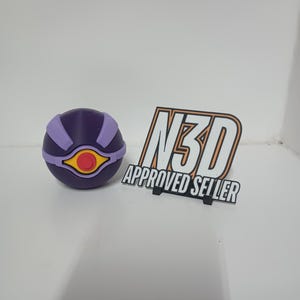 Puede incluir: Un objeto esférico morado y lila claro con un diseño en forma de ojo amarillo y rojo. Junto a él, un letrero en blanco y negro que dice "N3D APPROVED SELLER".