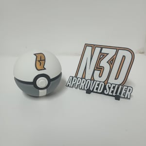 Puede incluir: Una bola blanca y gris con un botón central negro y blanco y un emblema dorado. Junto a ella hay un letrero que dice "N3D APPROVED SELLER" en negro y naranja.