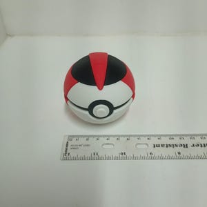 May include: A red, black, and white toy ball with a button in the center. The ball is approximately 2 inches in diameter. The toy is on a white surface with a ruler in the background.