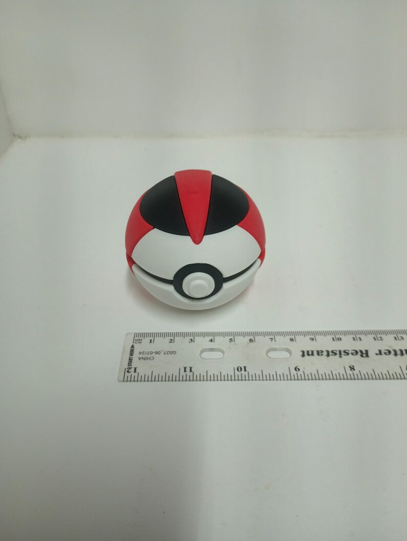 May include: A red, black, and white toy ball with a button in the center. The ball is approximately 2 inches in diameter. The toy is on a white surface with a ruler in the background.