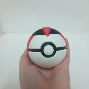 May include: A white, red, and black Pok&eacute; Ball toy. The ball is held in a hand and has a circular design with a black center and a white button. The top and sides are red and black.