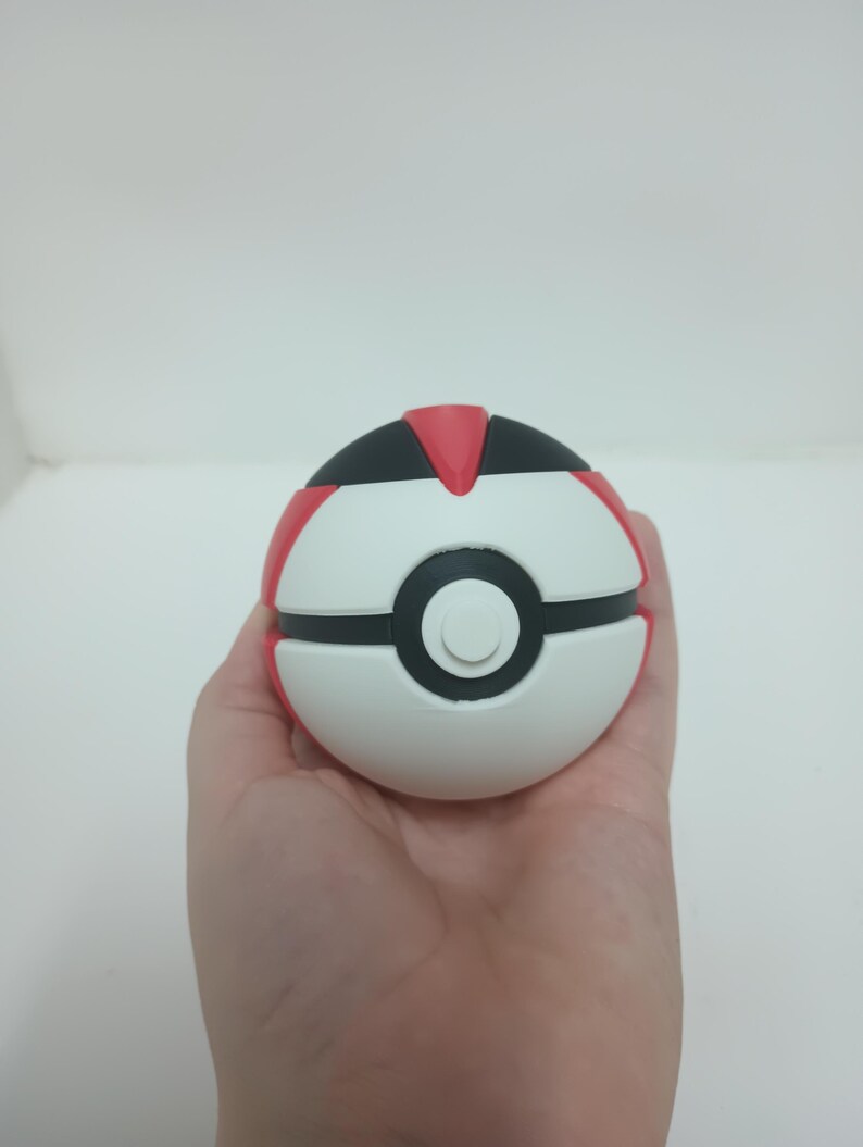 May include: A white, red, and black Pok&eacute; Ball toy. The ball is held in a hand and has a circular design with a black center and a white button. The top and sides are red and black.