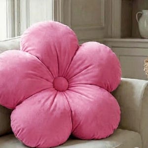 Puede incluir: Un gran cojín decorativo rosa con forma de flor descansa sobre un sofá beige. El cojín tiene cinco pétalos redondeados y un núcleo central circular. El fondo incluye una ventana, un mueble y un jarrón con flores secas.