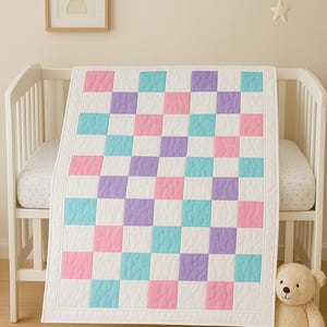 May include: A baby quilt with a checkerboard pattern in pastel colors of pink, blue, and purple. The quilt is draped over a white crib. A framed bunny art print hangs on the wall, and a stuffed teddy bear sits on the floor.