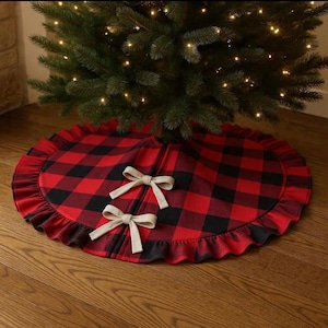 May include: A red and black buffalo plaid Christmas tree skirt with a ruffled edge and two beige bows. The skirt is circular and sits beneath a green Christmas tree with twinkling lights, creating a festive holiday display.