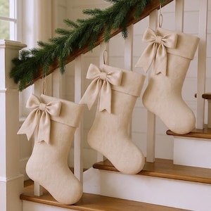 Op de afbeelding: Drie beige kerstsokken met bijpassende strikken hangen aan een witte trapleuning versierd met groenblijvende slingers. De sokken zijn gemaakt van een zachte stof en hebben een omgeslagen manchet.