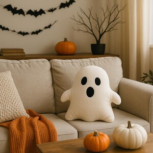 May include: A cream ghost-shaped cushion with black eyes and mouth rests on a beige sofa. The scene includes pumpkins, a bat garland, and a decorative tree, creating a Halloween atmosphere.