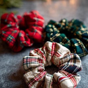 Puede incluir: Tres coleteros a cuadros en colores rojo, verde y crema. Los coleteros están hechos de tela con un patrón de cuadros clásico, perfectos para añadir un toque de estilo a cualquier peinado. Los coleteros están sobre una superficie gris.